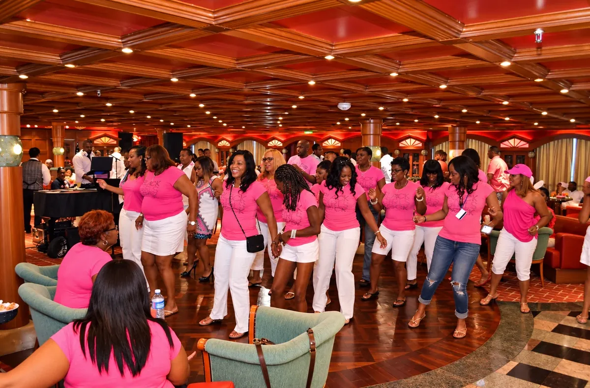 Guests in coordinated pink tops and white outfits line dancing across a cruise lounge floor
